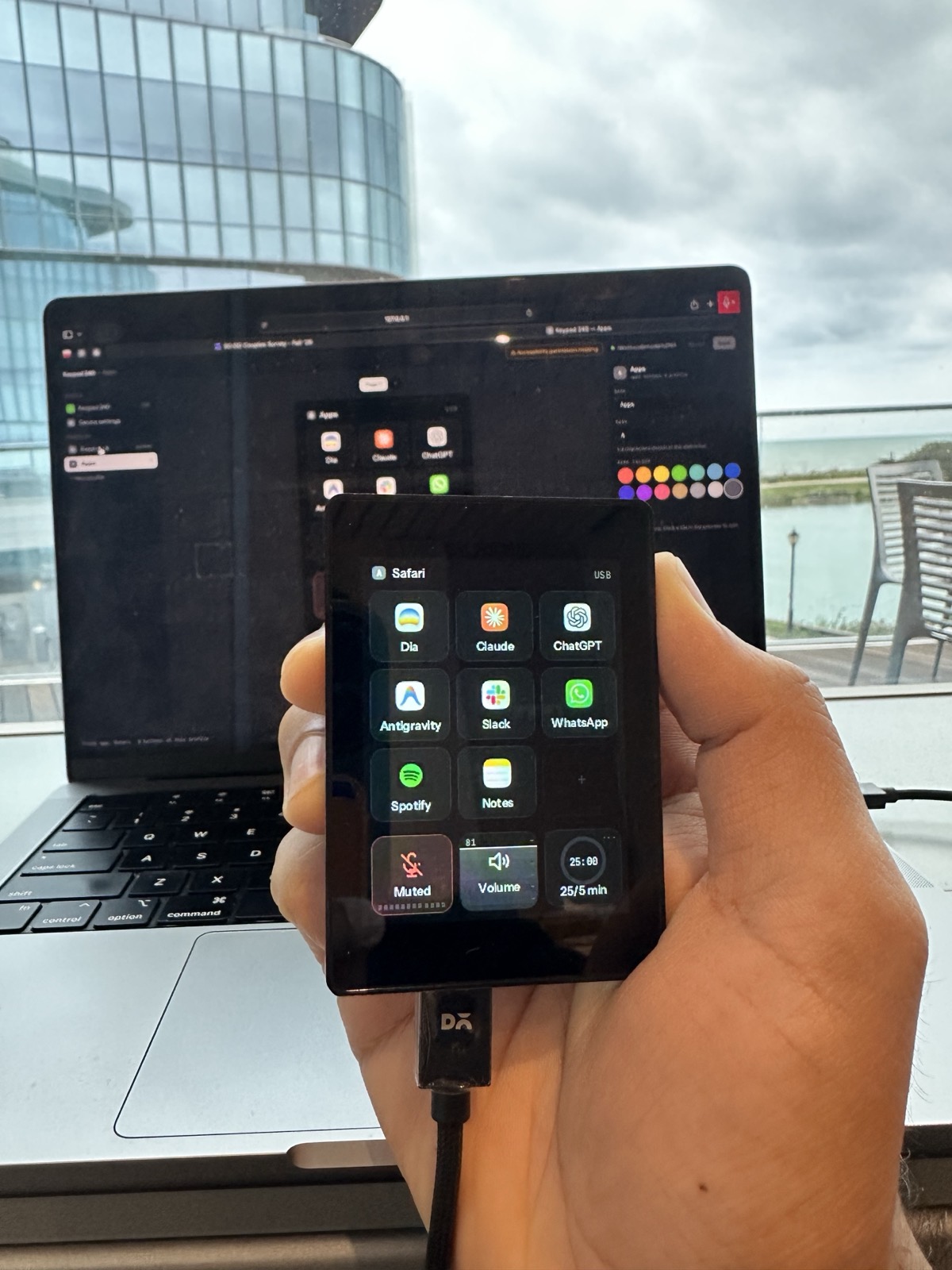 QuickPad held up in front of a MacBook, showing eight app launchers above mic, volume and focus timer tiles